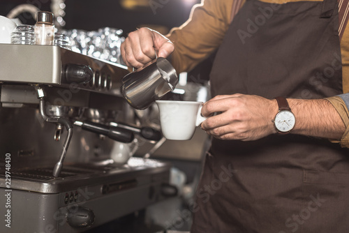 cropped shot of barista in apron pouring milk into coffee while preparing it in restaurant