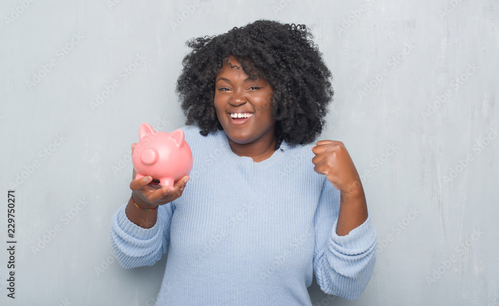 Φωτογραφία Young african american woman over grey grunge wall holding piggy bank screaming