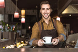 smiling young barista holding cup of coffee and looking at camera