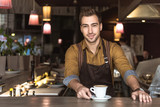 attractive young barista with cup of delicious coffee looking at camera