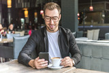 smiling young man with cup of coffee using smartphone in cafe
