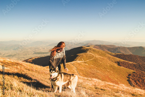 Red-hair girl in blue sweater with ornament play with husky. Ukrainian Carpathians in autumn time. Warm colored green leaves.Forests and mountains  top view. Travel with dog