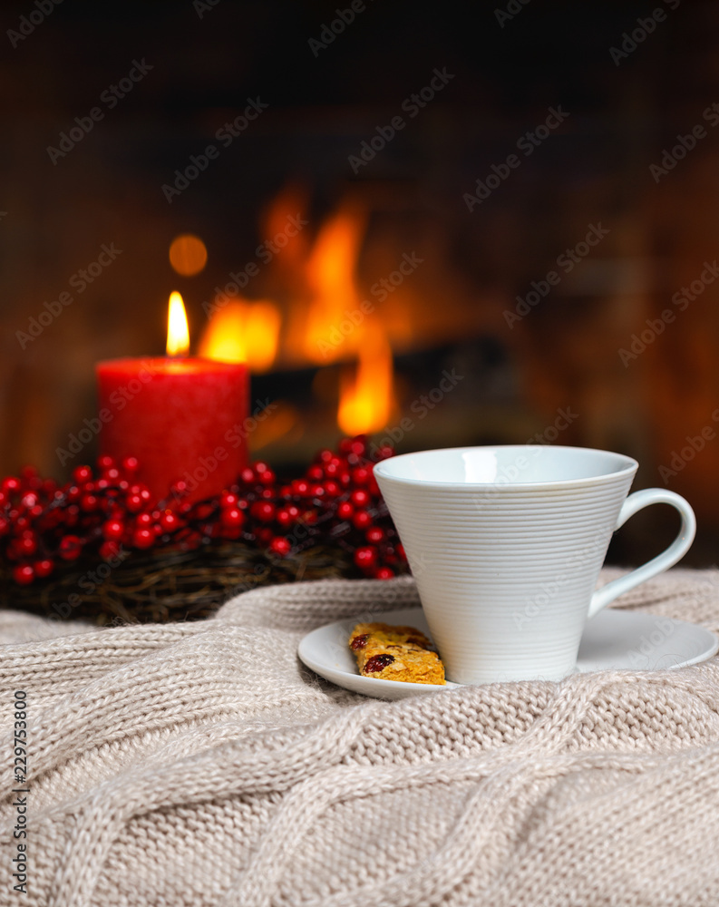 Cup of hot drink with cookie berries and red candle in red Christmas decoration on cozy knitted plaid in front of fireplace. Christmas New Year concept. Cozy relaxed magical atmosphere home interior. Cup of hot drink with cookie berries and red candle in red Christmas decoration on cozy knitted plaid in front of fireplace. Christmas New Year concept. Cozy relaxed magical atmosphere home interior.