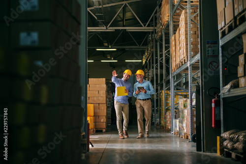 Two successful smiling business man walking through big warehouse with helmets on their heads.Younger man is shoving older one shelf’s full of products ready to be delivered. Happy investors.