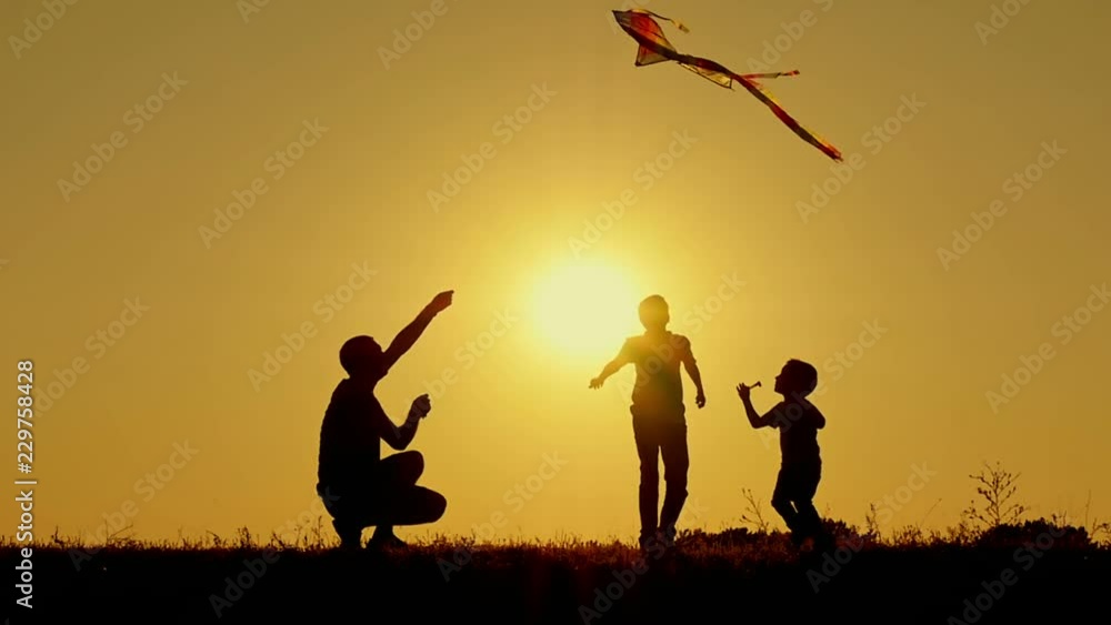 A happy father with two children playing in the open air running a flying kite. Silhouette of unrecognizable people at sunset. A happy father with two children playing in the open air running a flying kite. Silhouette of unrecognizable people at sunset.