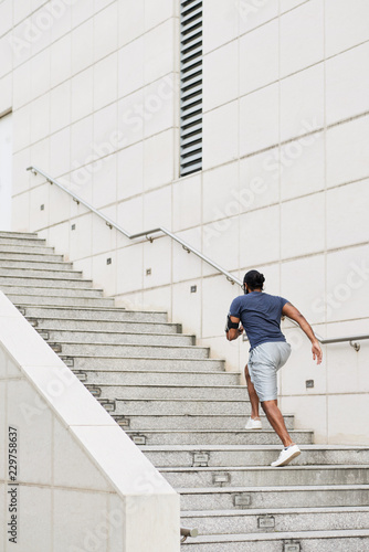 Rear view of unrecognizable sportsman with smartphone on shoulder running up concrete steps while having morning workout