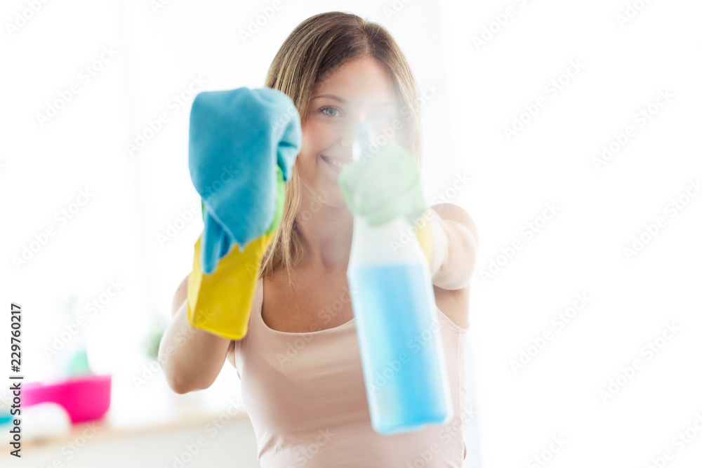 Beautiful young woman using a duster and a spray and smiling while cleaning windows at home. Beautiful young woman using a duster and a spray and smiling while cleaning windows at home.