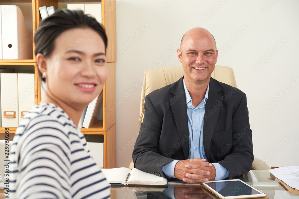 Portrait of confident smiling businessman sitting at his workplace with young woman, she came at meeting Portrait of confident smiling businessman sitting at his workplace with young woman, she came at meeting