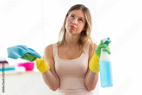 Boring young woman holding a duster and spray at home.