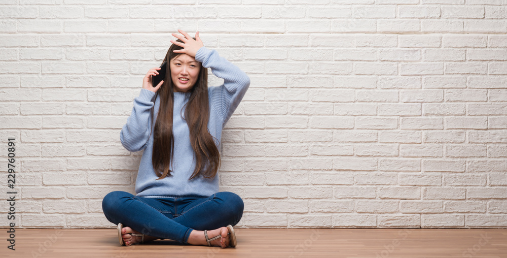 Young Chinese woman sitting on the floor over brick wall stressed with hand on head, shocked with shame and surprise face, angry and frustrated. Fear and upset for mistake. Young Chinese woman sitting on the floor over brick wall stressed with hand on head, shocked with shame and surprise face, angry and frustrated. Fear and upset for mistake.