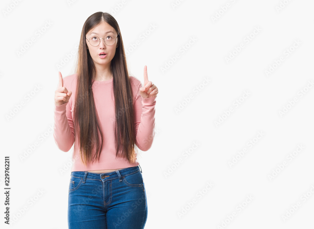 Young Chinese woman over isolated background wearing glasses amazed and surprised looking up and pointing with fingers and raised arms. Young Chinese woman over isolated background wearing glasses amazed and surprised looking up and pointing with fingers and raised arms.