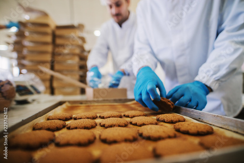 Close up of tray full of fresh baked cookies in food factory. Blurred picture of two male employees in sterile clothes packing cookies in background.