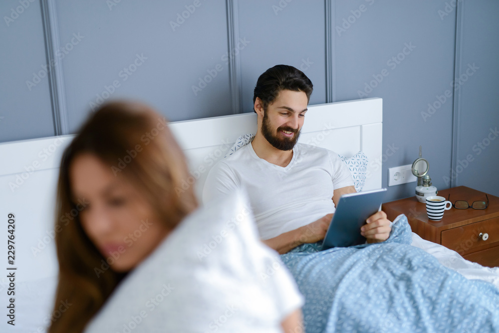 Beautiful young girl sitting on bed and looking sad while hers boyfriends do not pay attention to her. He is smiling and looking at his tablet.
