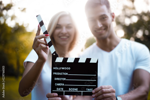 Cute loving couple standing on grass in nature green park with beautiful sundown light. Looking camera holding cinema film making clapperboard.