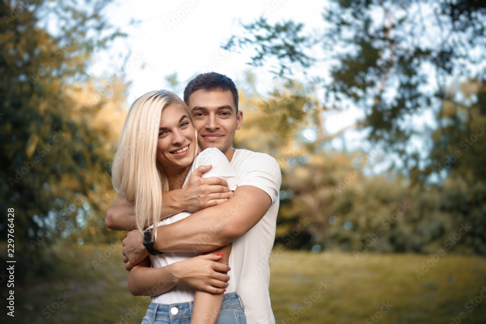 Cute loving couple standing on grass in nature green park with beautiful sundown light. Looking camera hugging. Cute loving couple standing on grass in nature green park with beautiful sundown light. Looking camera hugging.