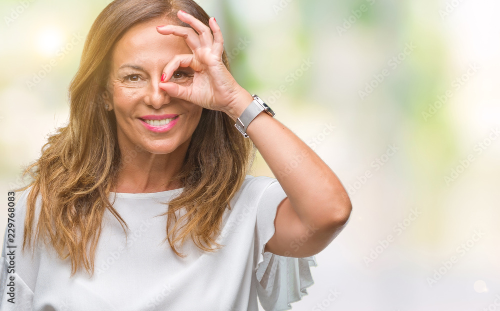 Middle age senior hispanic woman over isolated background doing ok gesture with hand smiling, eye looking through fingers with happy face.