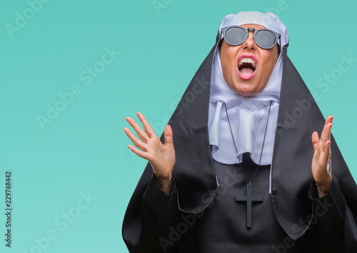 Konstfotografi Middle age senior catholic nun woman wearing sunglasses over isolated background crazy and mad shouting and yelling with aggressive expression and arms raised