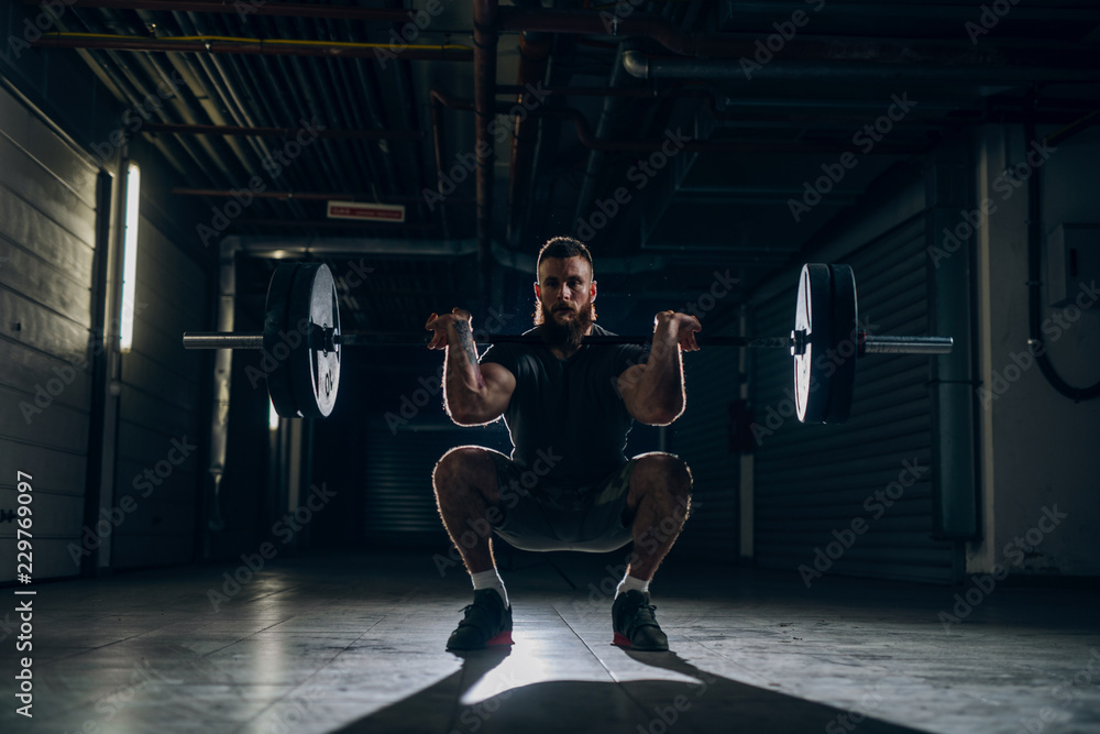 Muscular attractive caucasian bearded man doing front barbell squat in underground hallway. Storage units and pipes in background. Muscular attractive caucasian bearded man doing front barbell squat in underground hallway. Storage units and pipes in background.