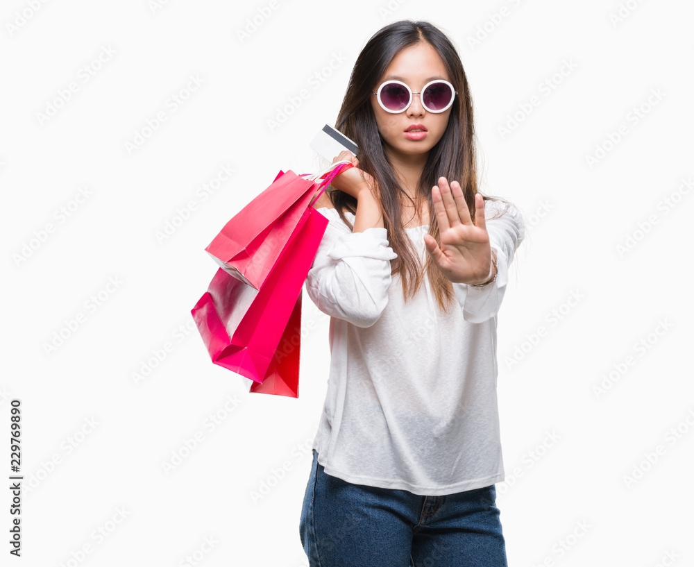Young asian woman holding shopping bags on sales over isolated background with open hand doing stop sign with serious and confident expression, defense gesture Young asian woman holding shopping bags on sales over isolated background with open hand doing stop sign with serious and confident expression, defense gesture