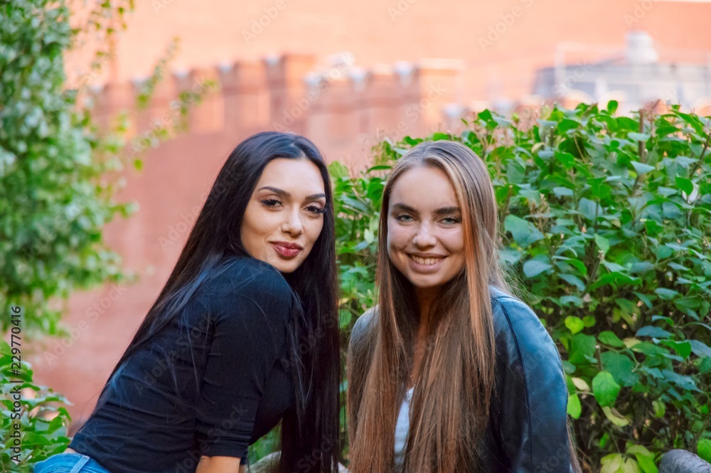 Girlfriend girls laugh in the Park on the background of green bushes and a red wall. Two girls in the park
