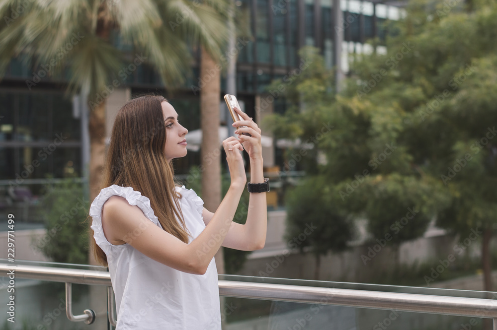 Young european female with brown hair and in white casual style blouse taking photos with her mobile phone. Woman taking a selfie outdoors Young european female with brown hair and in white casual style blouse taking photos with her mobile phone. Woman taking a selfie outdoors