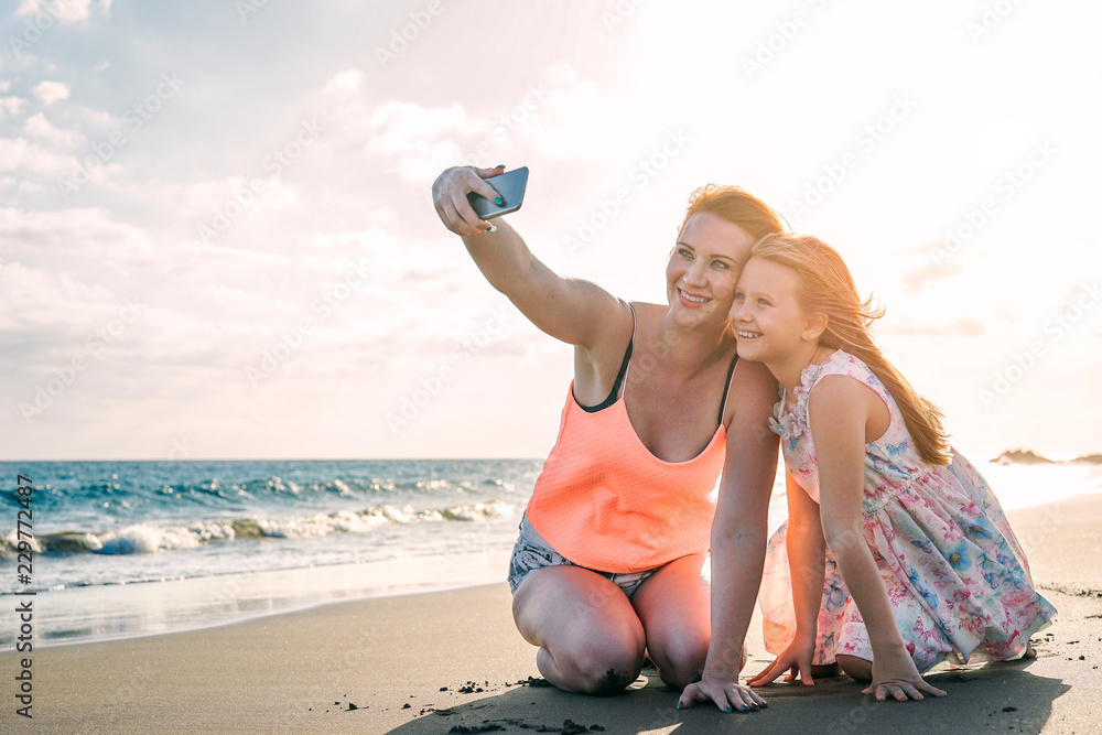Happy loving family mother and daughter taking a selfie with mobile smart phone on the beach at sunset - Mom with her kid in holiday - Parent, vacation, tech and family lifestyle concept Happy loving family mother and daughter taking a selfie with mobile smart phone on the beach at sunset - Mom with her kid in holiday - Parent, vacation, tech and family lifestyle concept