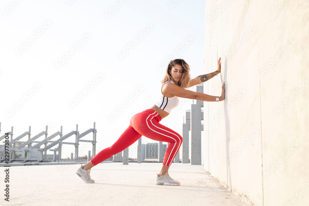 Beautiful caucasian athletic female with long legs wearing a bright red long tights does a stretch against a concrete wall with a open outdoor stadium in the background.  