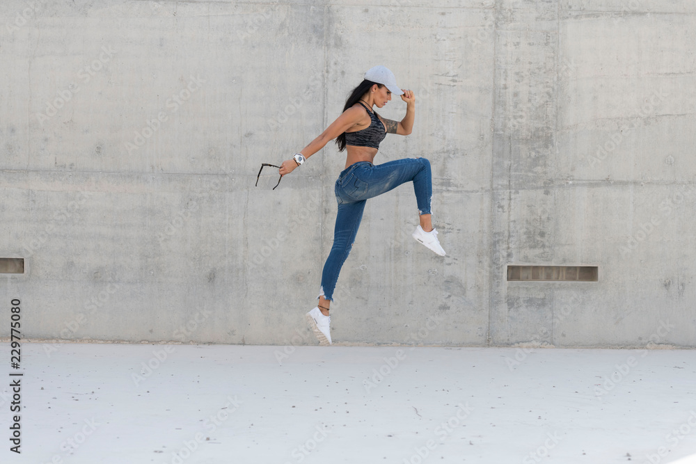 Beautiful caucasian athletic girl with long black hair wearing blue jeans and black sports bra and wearing a cap jumps   in a  concrete stadium  on a bright sunny day