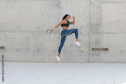 Beautiful caucasian athletic girl with long black hair wearing blue jeans and black sports bra and wearing a cap jumps in a concrete stadium on a bright sunny day