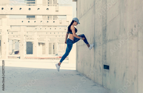 Beautiful caucasian athletic girl with long black hair wearing blue jeans and black sports bra and wearing a cap jumps in a concrete stadium on a bright sunny day