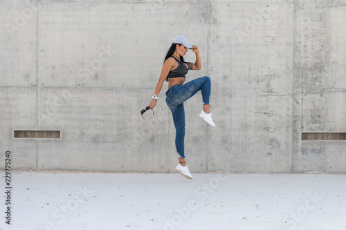 Beautiful caucasian athletic girl with long black hair wearing blue jeans and black sports bra and wearing a cap jumps in a concrete stadium on a bright sunny day © Paul