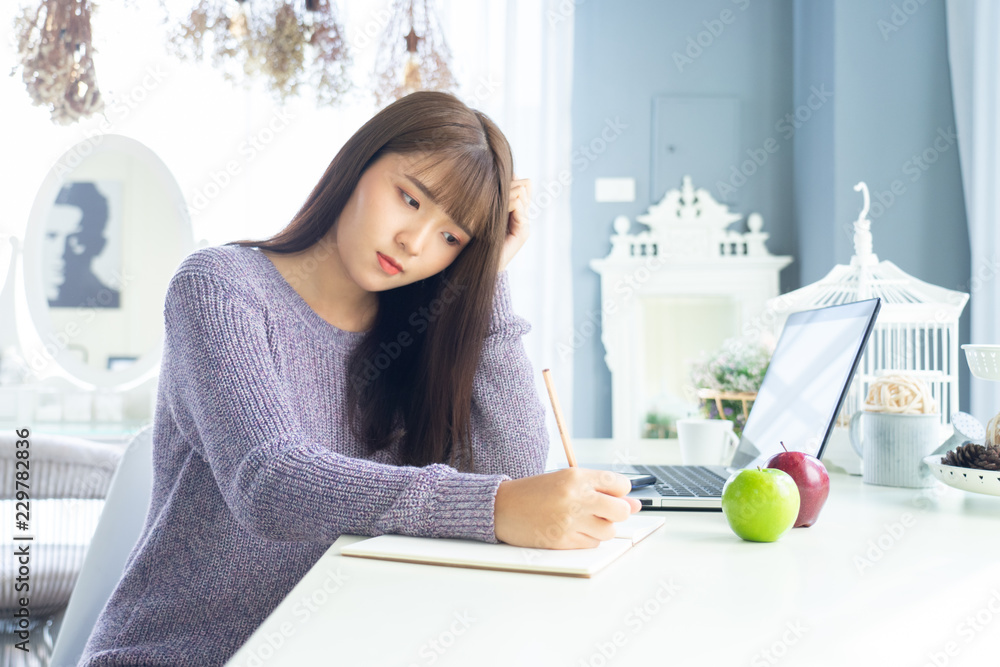 Happy asian beautiful woman working on a laptop sitting on the table in the house.Morning she dress sweater and work at home . Happy asian beautiful woman working on a laptop sitting on the table in the house.Morning she dress sweater and work at home .