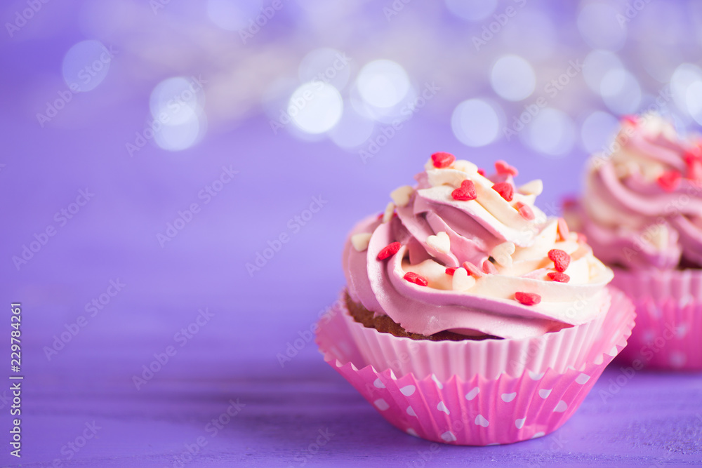 Closeup two cupcakes with creamy pink and white top decorated with little hearts on purple bokeh background. Closeup two cupcakes with creamy pink and white top decorated with little hearts on purple bokeh background.