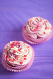 Closeup two cupcakes with creamy pink and white top decorated with little hearts on purple wooden background