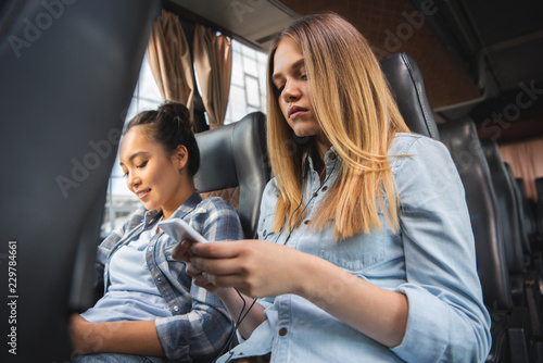 attractive woman sitting with smartphone while her asian female friend sitting near during trip on travel bus