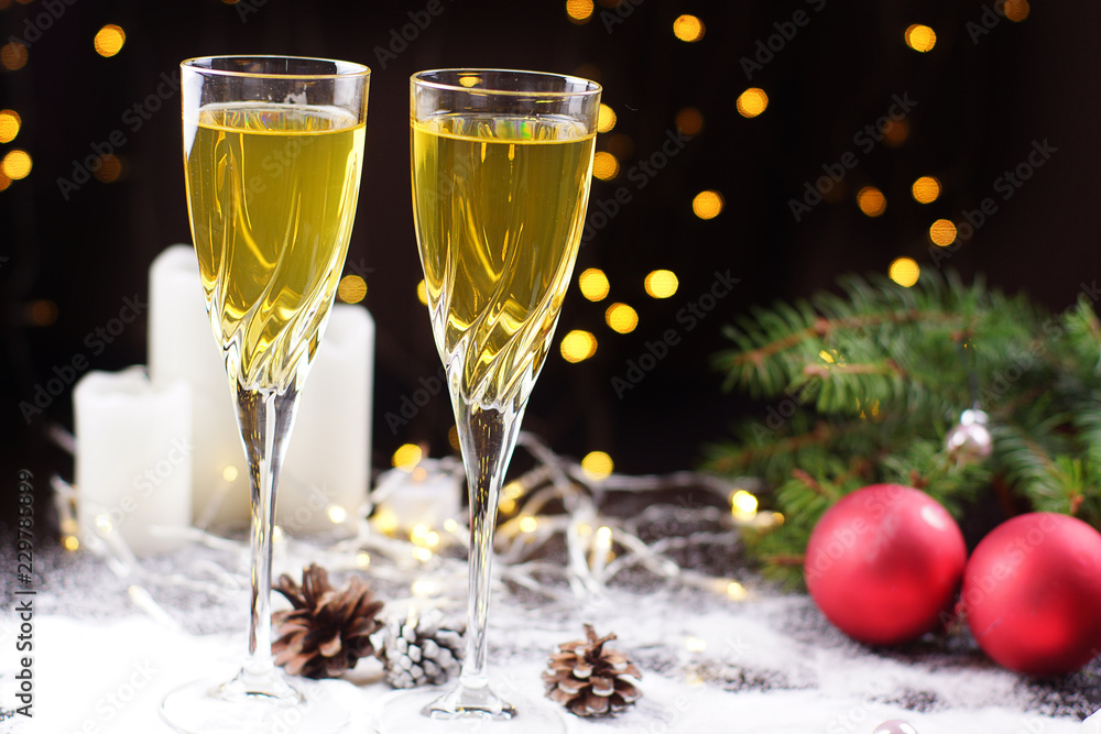 Two glasses of champagne or juice on the table, decorated with a glowing garland, red balls, a Christmas tree branch and white candles Two glasses of champagne or juice on the table, decorated with a glowing garland, red balls, a Christmas tree branch and white candles