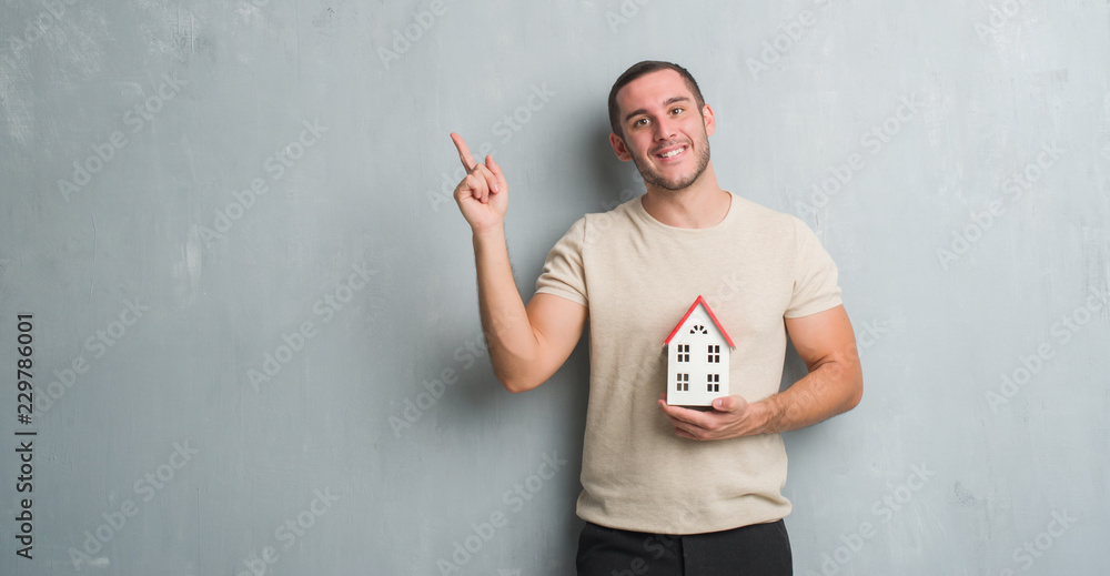 Young caucasian real state agent man over grey grunge wall holding house very happy pointing with hand and finger to the side Young caucasian real state agent man over grey grunge wall holding house very happy pointing with hand and finger to the side