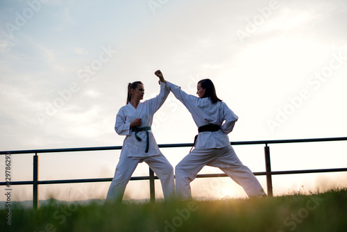 Two fit young women dressed in white kimono training karate martial arts in nature.