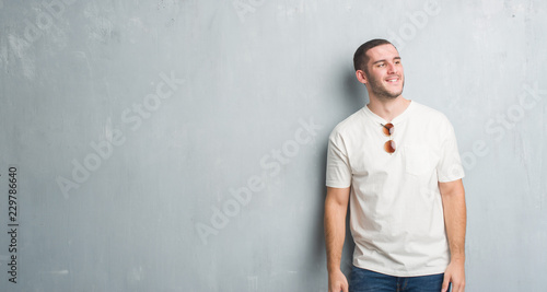 Young caucasian man over grey grunge wall wearing sunglasses looking away to side with smile on face, natural expression. Laughing confident.