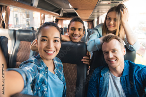 selective focus of smiling asian woman taking selfie with multicultural friends in travel bus