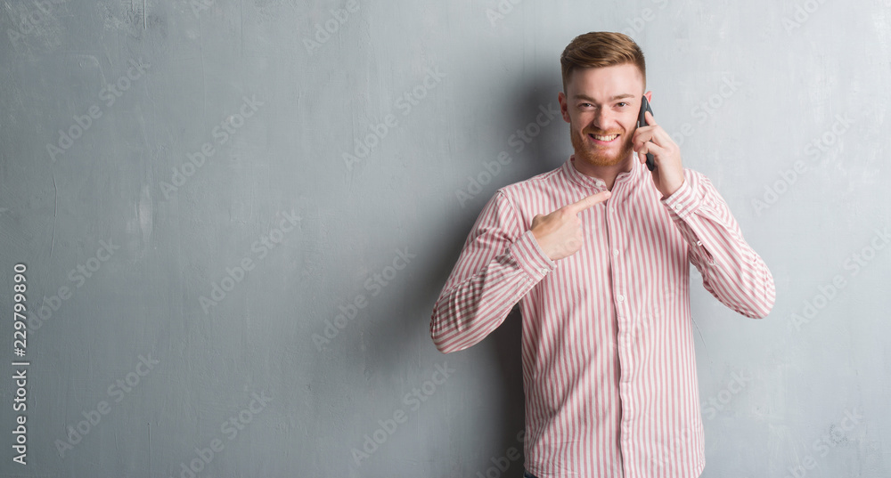 Young redhead man over grey grunge wall talking on the phone very happy pointing with hand and finger
