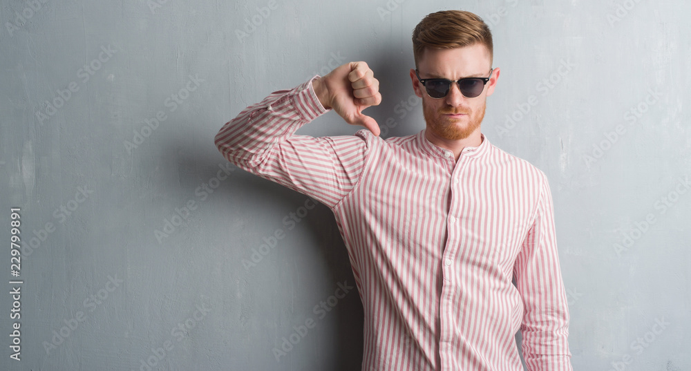 Young redhead man over grey grunge wall wearing sunglasses with angry face, negative sign showing dislike with thumbs down, rejection concept Young redhead man over grey grunge wall wearing sunglasses with angry face, negative sign showing dislike with thumbs down, rejection concept