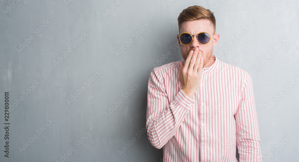 Young redhead man over grey grunge wall wearing retro sunglasses cover mouth with hand shocked with shame for mistake, expression of fear, scared in silence, secret concept Young redhead man over grey grunge wall wearing retro sunglasses cover mouth with hand shocked with shame for mistake, expression of fear, scared in silence, secret concept