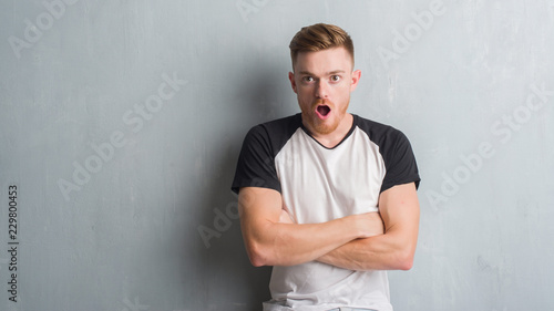 Young redhead man over grey grunge wall afraid and shocked with surprise expression, fear and excited face.