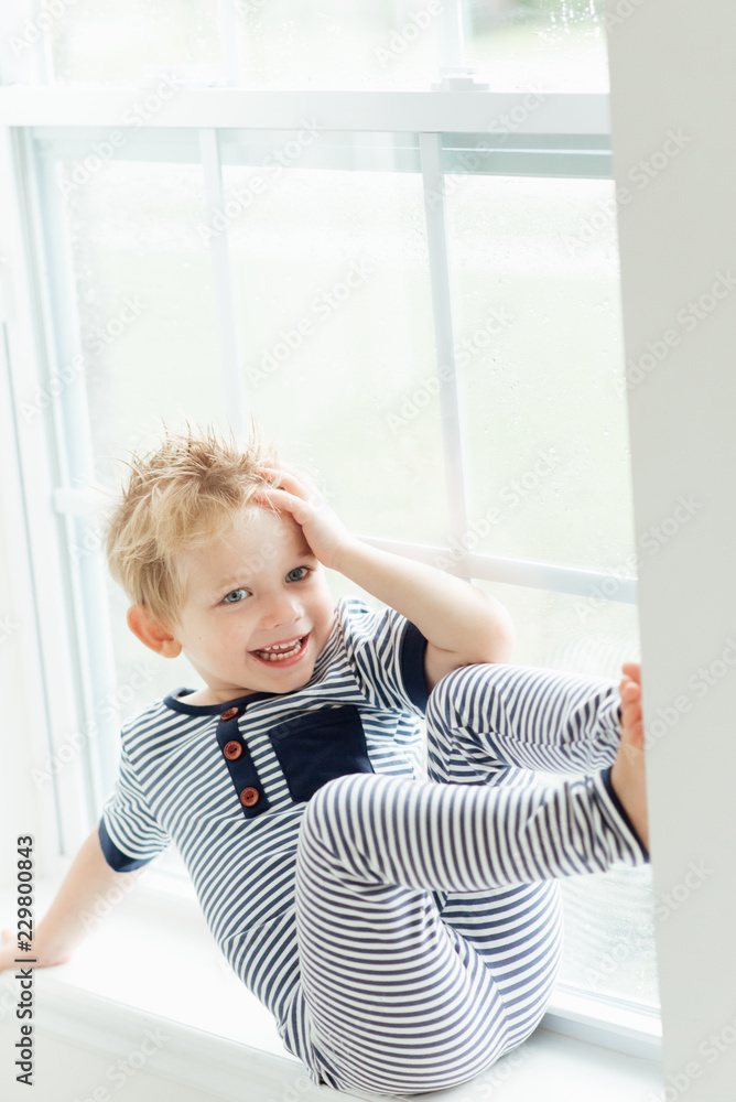 Smiling boy sitting on window sill with his hand on his head Smiling boy sitting on window sill with his hand on his head