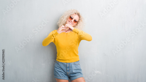 Young blonde woman with curly hair over grunge grey background smiling in love showing heart symbol and shape with hands. Romantic concept.