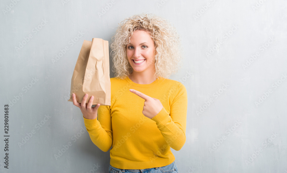 Young blonde woman over grunge grey background holding lunch paper bag very happy pointing with hand and finger Young blonde woman over grunge grey background holding lunch paper bag very happy pointing with hand and finger