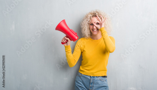 Young blonde woman over grunge grey background holding megaphone with happy face smiling doing ok sign with hand on eye looking through fingers