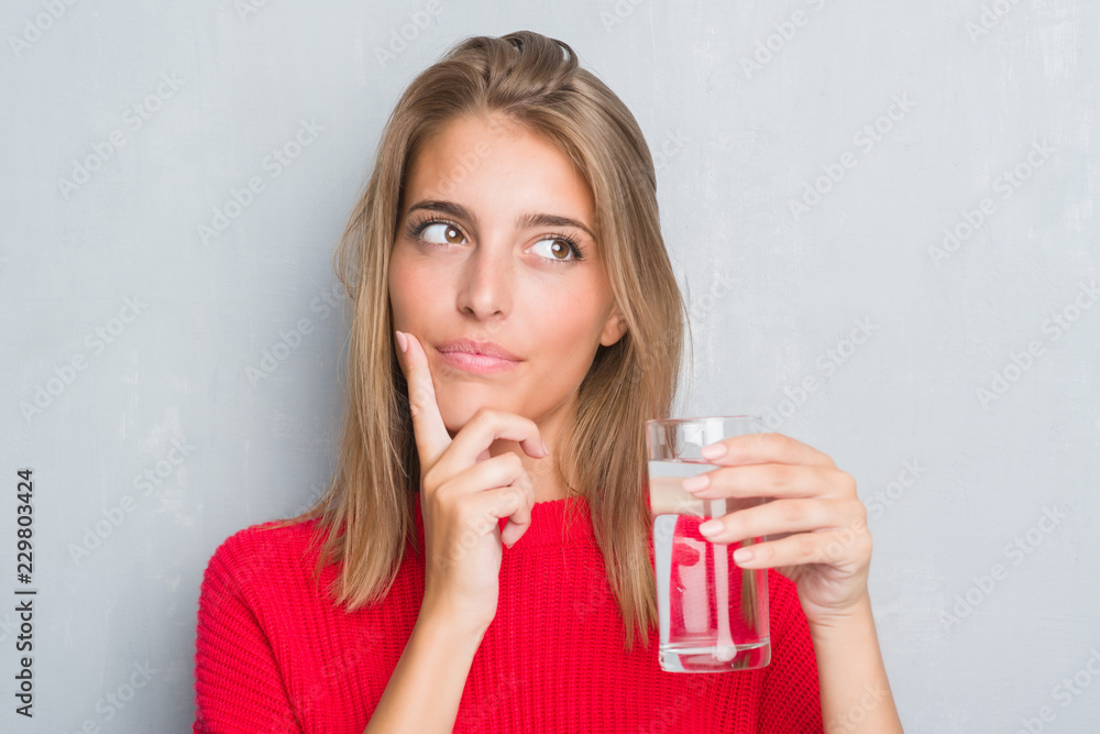 Beautiful young woman over grunge grey wall drinking a glass of water serious face thinking about question, very confused idea Beautiful young woman over grunge grey wall drinking a glass of water serious face thinking about question, very confused idea