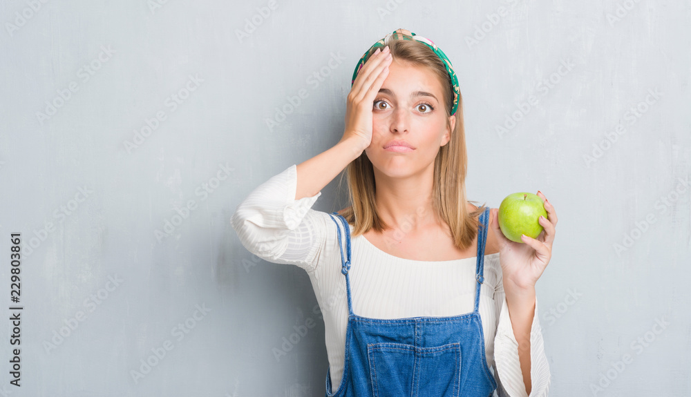 Beautiful young woman over grunge grey wall eating green apple stressed with hand on head, shocked with shame and surprise face, angry and frustrated. Fear and upset for mistake. Beautiful young woman over grunge grey wall eating green apple stressed with hand on head, shocked with shame and surprise face, angry and frustrated. Fear and upset for mistake.